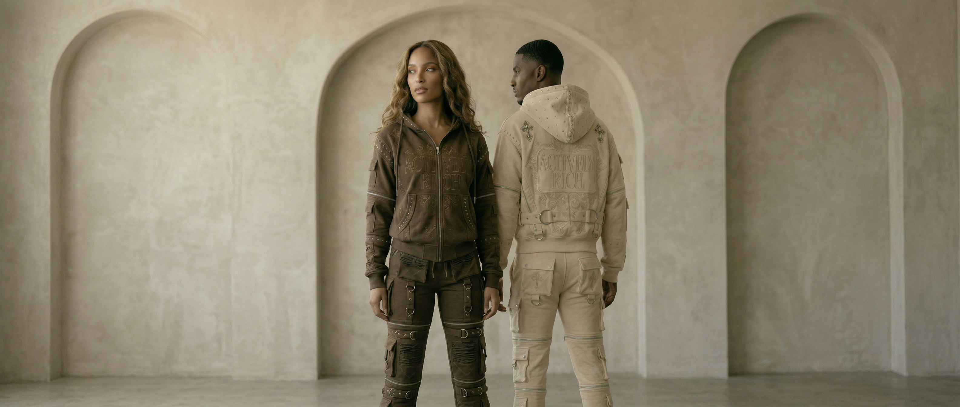 Actively Rich Ascension Noir luxury streetwear duo wearing brown and cream hoodie and cargo sets displayed in a wide neutral archway setting